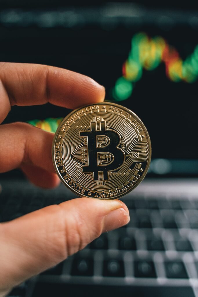 pexels-photo-6770513-6770513 Close-up of a hand holding a Bitcoin coin in front of a trading screen, symbolizing cryptocurrency trading.